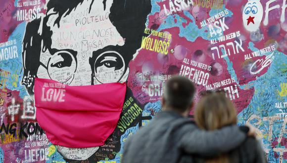 FILE - A couple look at the "Lennon Wall" with a face mask attached to the image of John Lennon, in Prague, Czech Republic, on April 6, 2020. Like so many other events in the year of coronavirus, an annual tribute to John Lennon held in its adopted city of New York will go online. The five-hour event will be streamed for free on Lennon's birthday, October 9, starting at 7 p.m. Eastern time on the LennonTribute.org website. It will feature recorded performances from Patti Smith, Rosanne Cash, Natalie Merchant, Jackson Browne, Jorma Kaukonen and others. (AP Photo/Petr David Josek)