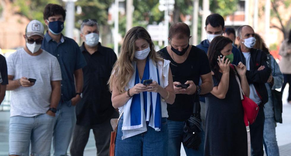 Una mujer con una bandera uruguaya al hombro hace fila con otras para ser inoculadas con la vacuna CoronaVac en el Antel Arena de Montevideo, el 29 de marzo de 2021. (Pablo PORCIUNCULA / AFP).
